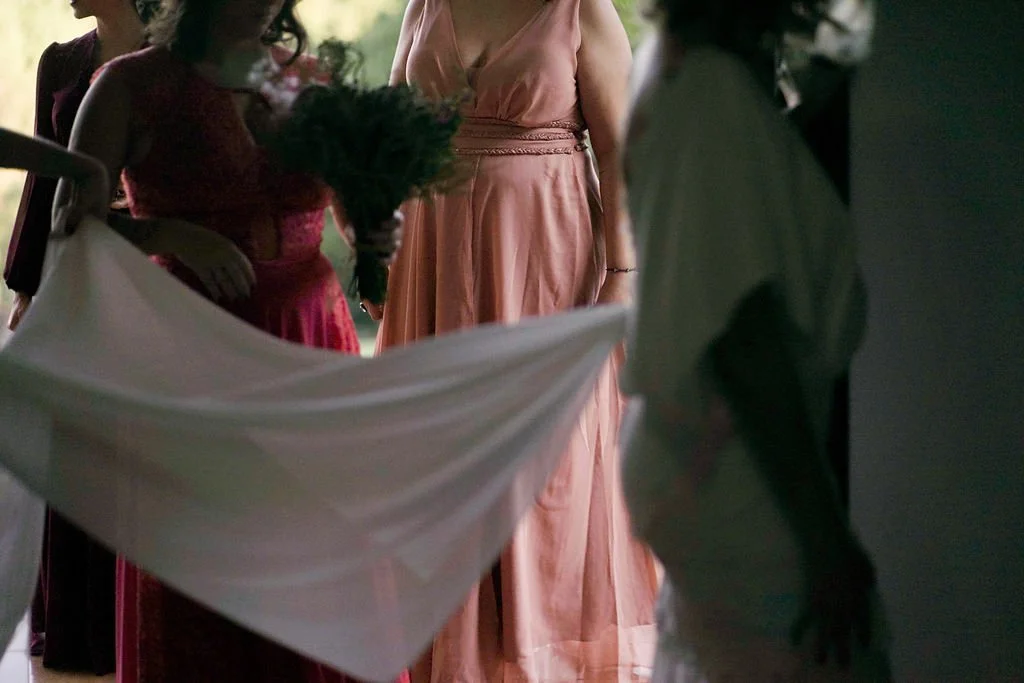 Mulher com vestido rosa claro e outras pessoas ao redor em um evento, com decoração de tecido branco suspenso.