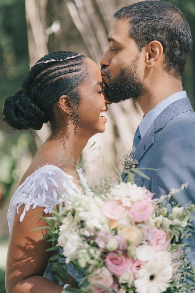 Casal de noivos se beijando, segurando um buquê de flores, ao ar livre.