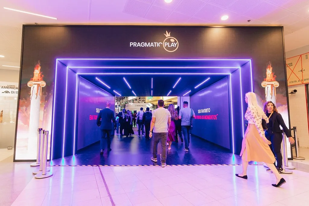 Entrada com iluminação de luzes neon azul, com o nome 'PRAGMATIC PLAY' acima, onde várias pessoas estão entrando em uma área de evento ou espaço de entretenimento dentro de um shopping ou centro comercial.