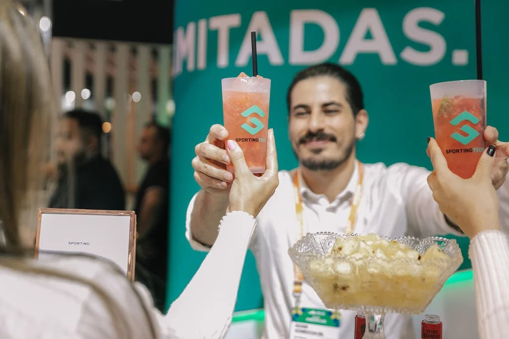 Homens celebrando com drinks em uma feira ou evento, com um jovem de cabelo escuro, barba e camisa branca sorrindo ao fundo. Pessoas ao redor e uma placa com o texto 'MITADAS.' ao fundo.