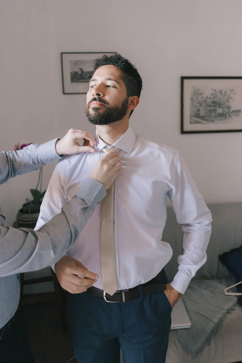 Homem na verdade, ajustando a gravata enquanto alguém o ajuda, em um ambiente interno decorado com quadros na parede.