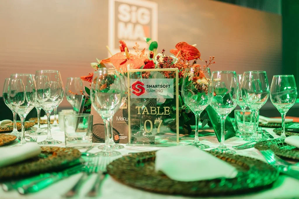 Mesa de jantar decorada com taças de vidro, flores e conchas, com sinalizador de mesa com texto 'TABLE 07' e logotipo da SmartSoft Gaming ao fundo.