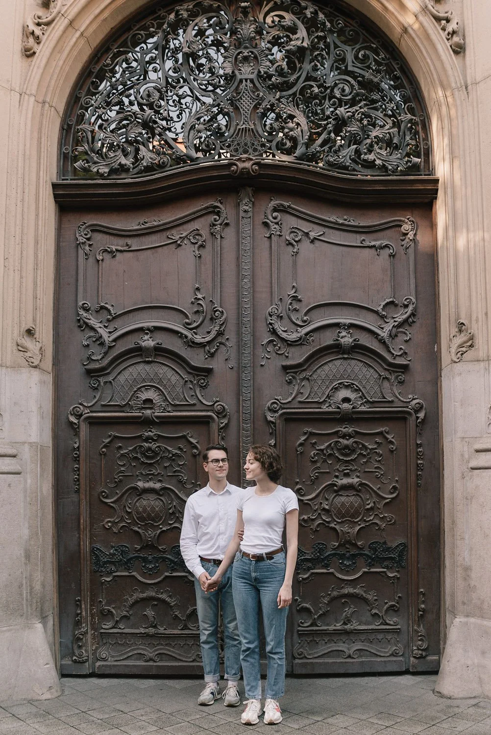 Homem e mulher de cabelo curto, usando roupas casual, segurando as mãos na frente de uma porta de ferro ornamentada.