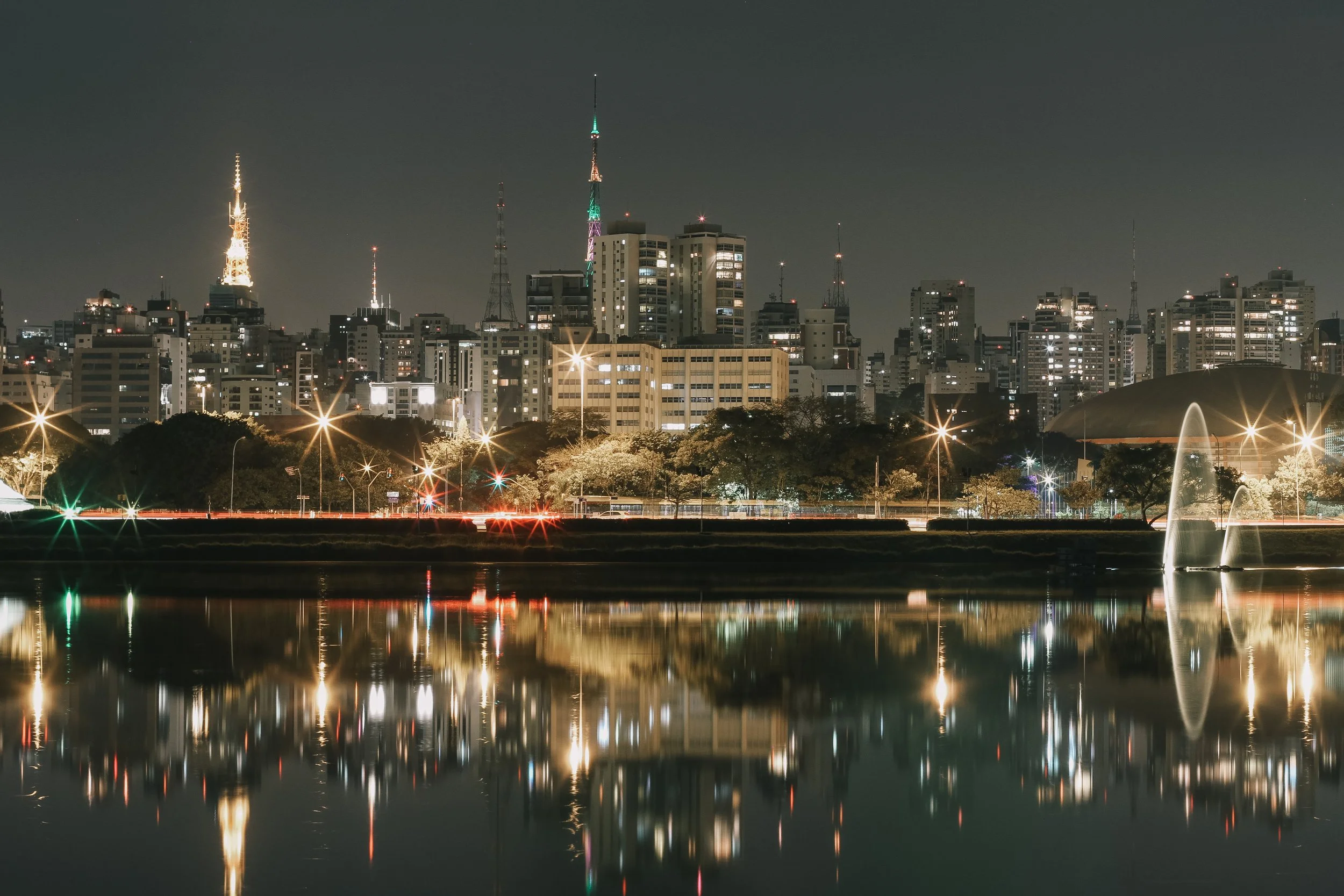 Cidade à noite vista do rio, com edifícios iluminados e reflexo na água, incluindo torres de transmissão com luzes coloridas e fonte no parque ao lado.