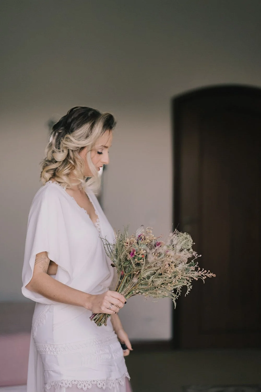 Mulher com cabelo cacheado de olhos fechados segurando um buquê de flores secas, usando roupa branca com detalhes de renda.