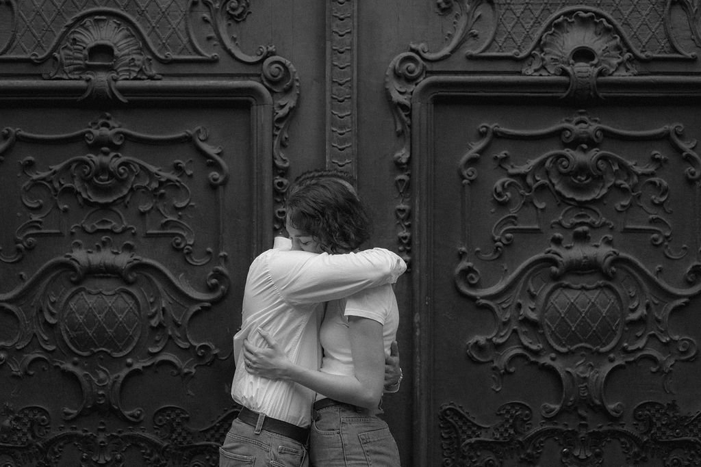 Dois jovens se abraçando na frente de uma porta de ferro decorada, em preto e branco.