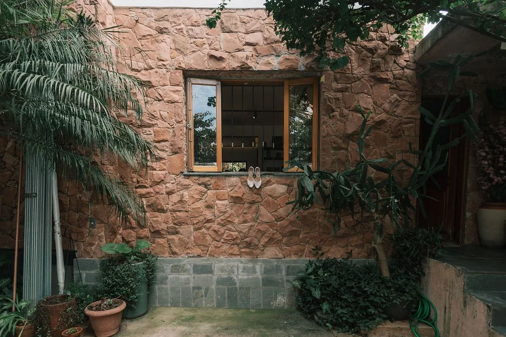 Parede de pedra, janela com vista para o interior, plantas ao redor, chinelos pendurados na janela