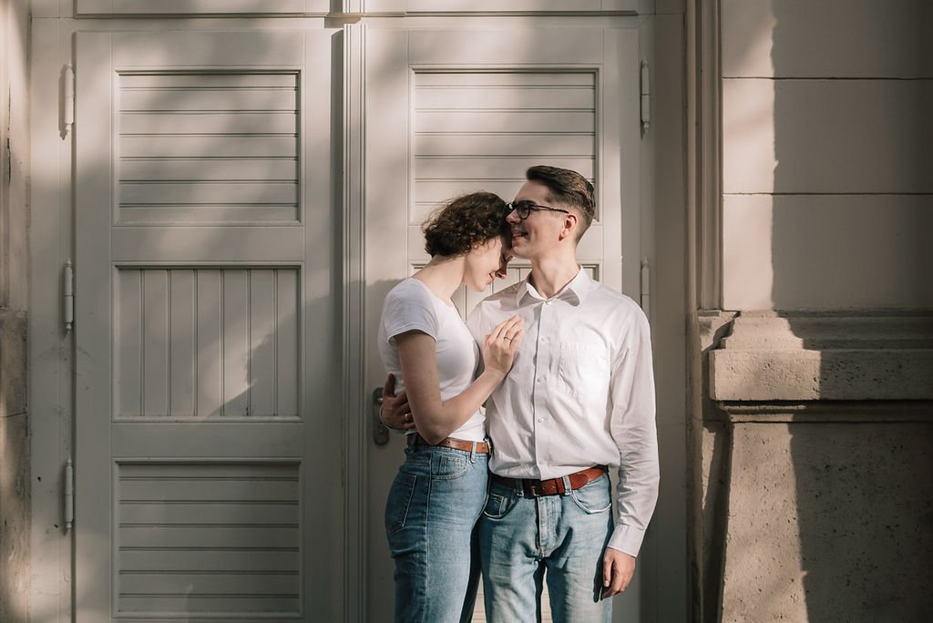Casal jovem se abraçando e sorrindo em frente a uma porta de madeira branca."}