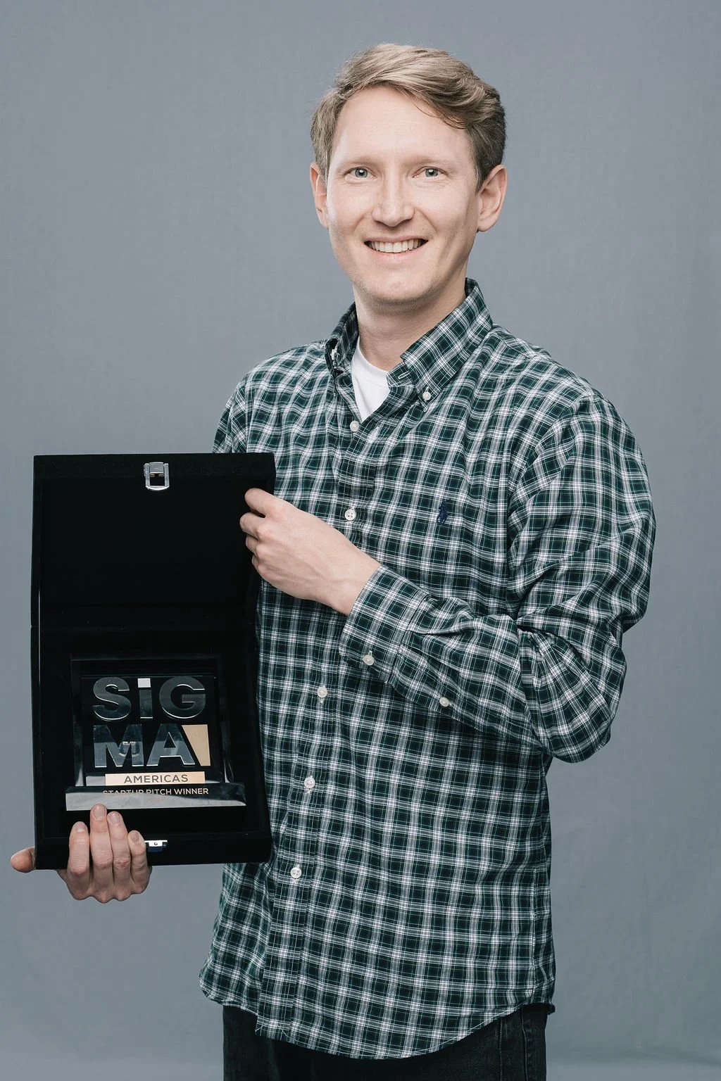 Homem sorridente segurando um troféu que diz 'SIGMA AMERICAS STARTUP PITCH WINNER'.