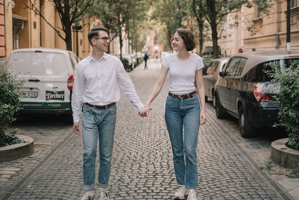 Um casal de jovens caminhando de mãos dadas na rua de paralelepípedo, com árvores e carros estacionados ao lado.