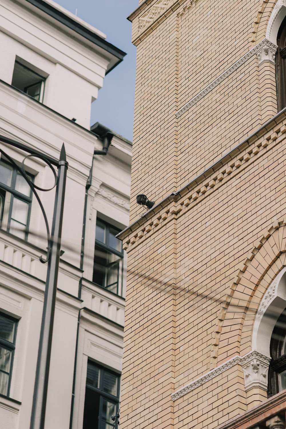 Imagem que mostra duas fachadas de prédios: uma branca com detalhes clássicos e uma de tijolos beges com ornamentos, com uma câmera de segurança instalada na parede de tijolos.