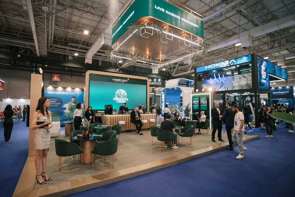 Ambiente de feira de negócios com vários estandes, pessoas conversando e sentadas, destaque para o estande do Dragon Tiger com tela grande e logo, e um palco com banner do provedor de cassino ao vivo.