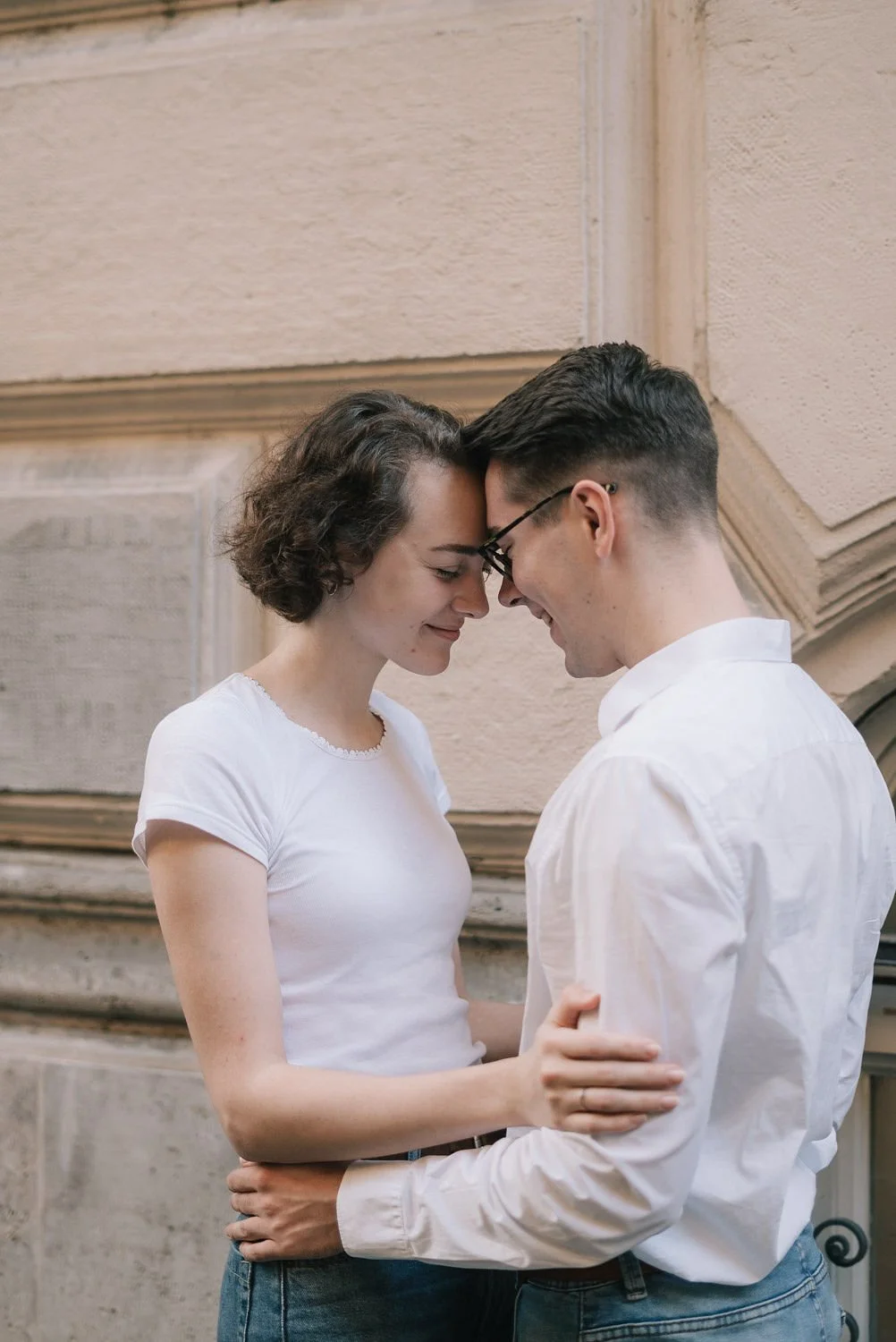 Casal se abraçando e tocando a testa, sorrindo com os olhos fechados, em um ambiente interno com parede de pedra ao fundo.