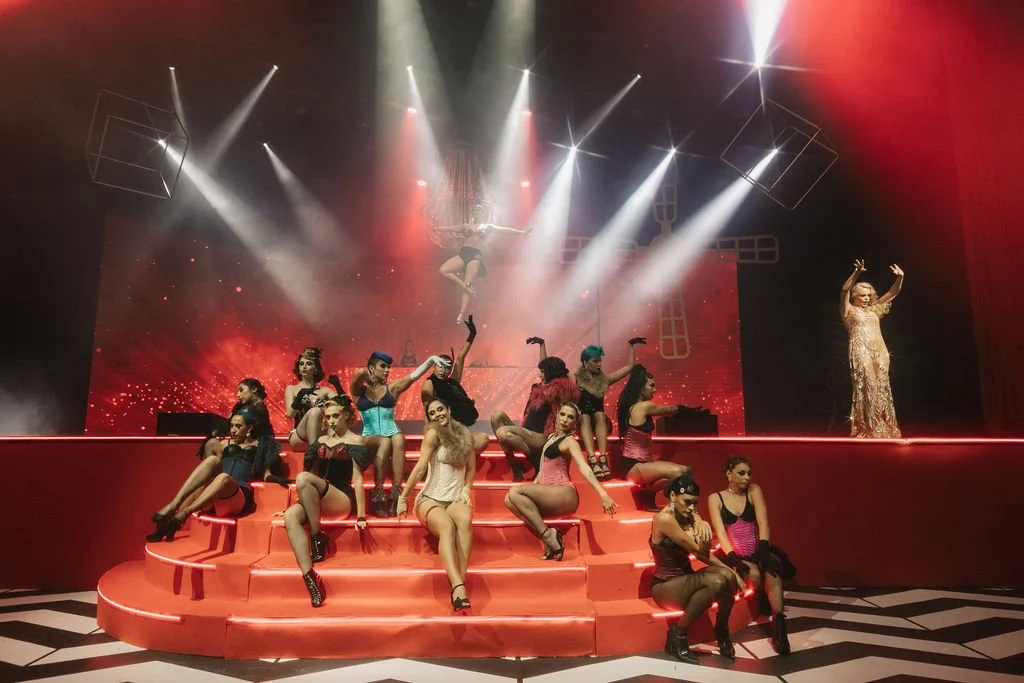 Grupo de performers em um palco desenhado com degraus vermelhos, com iluminação dramática e fundo vermelho, algumas usando roupas coloridas e roupas de veludo pretas, enquanto uma mulher com vestido dourado se apresenta ao lado.