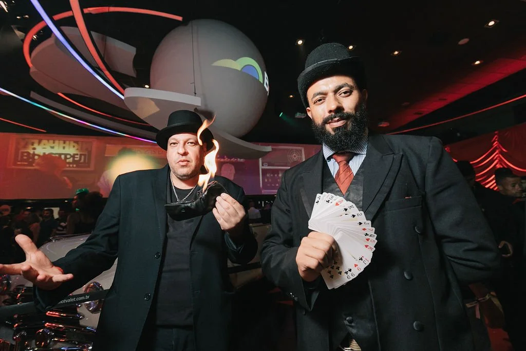 Dois homens com roupas formais e chapéus pretos estão em um ambiente com iluminação colorida. Um deles está segurando uma maleta em chamas, enquanto o outro exibe um baralho de cartas com uma expressão confiante.