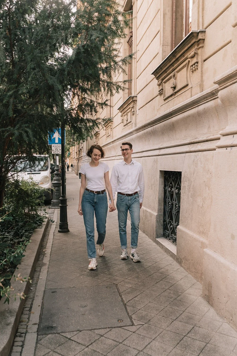 Casal jovem caminhando de mãos dadas na calçada de uma rua arborizada, com edifício antigo ao fundo e sinalização de bicicleta e direção.