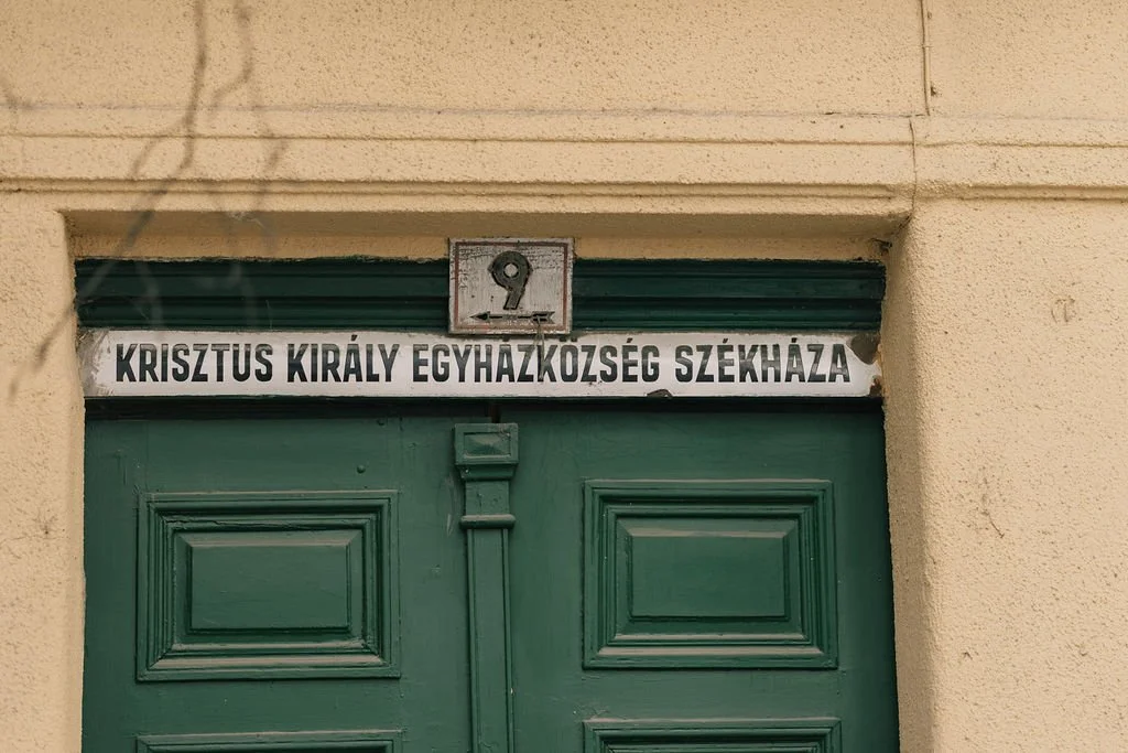Porta verde com placa acima que diz "KRISZTUS KIRÁLY EGYHÁZKOZÉSEG SZÉKHÁZA" e número 9 acima.