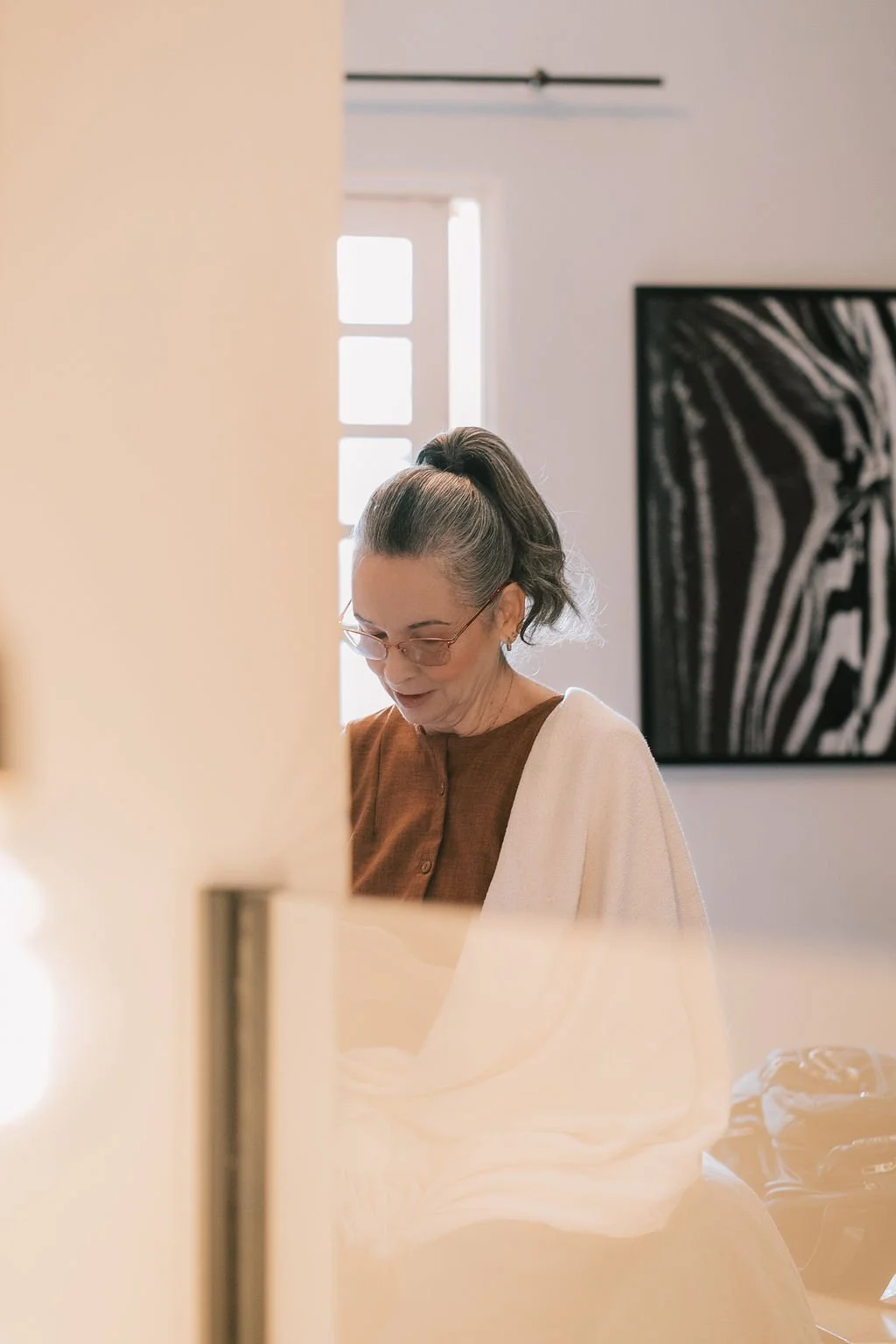 Mulher idosa de cabelos grisalhos presos em um coque, usando óculos e roupa marrom, de frente, em um ambiente com fundo artístico e uma janela com luz natural.