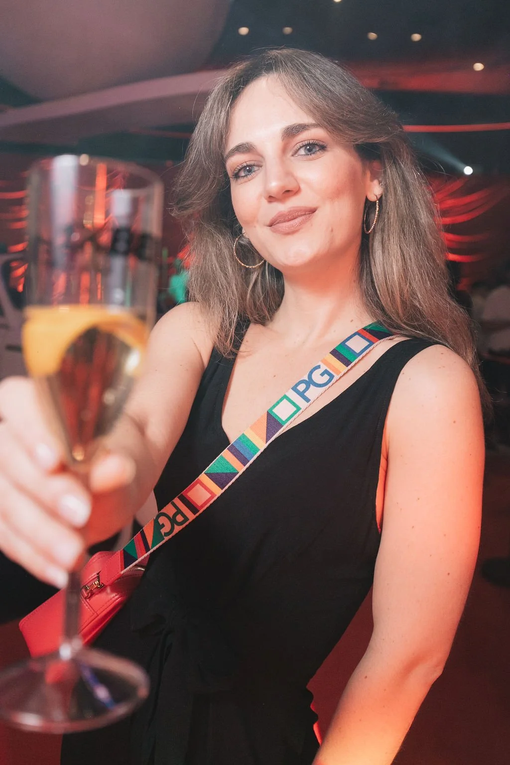 Mulher sorridente segurando uma taça de champanhe em um ambiente de festa ou evento, com iluminação colorida ao fundo.