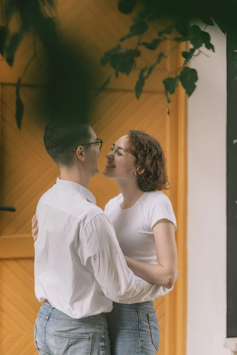 Um casal jovem, sorrindo, se abraçando, em um ambiente interno com parede de madeira ao fundo e plantas penduradas.