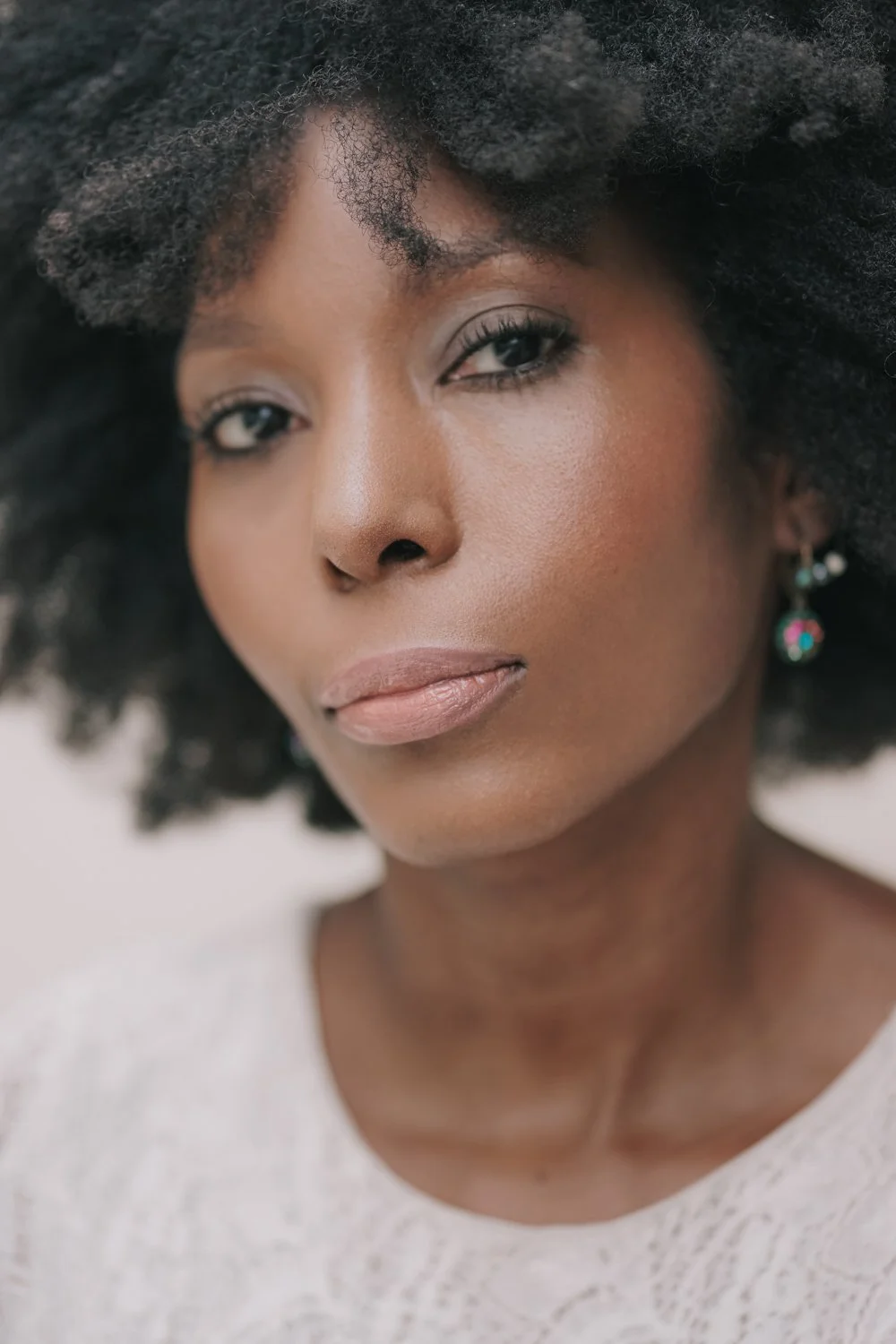 Retrato de uma mulher negra com cabelo afro, pele clara, usando maquiagem suave e brincos coloridos, com expressão tranquila