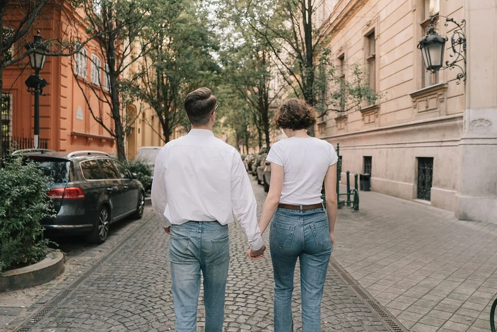 Casal caminhando de mãos dadas pela rua de paralelepípedos em uma cidade, com carros estacionados ao lado e edifícios históricos ao redor.