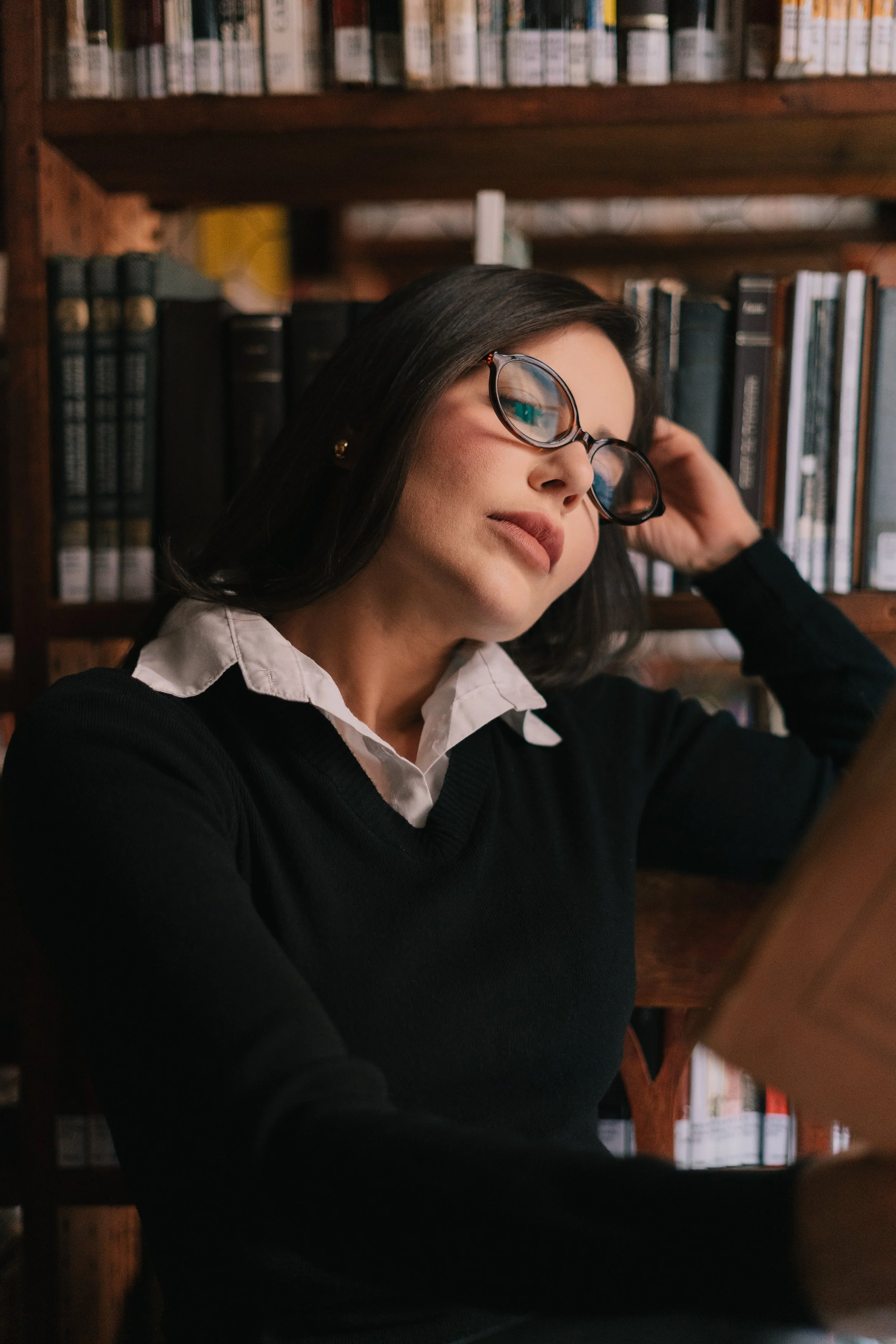 Mulher de óculos escuros, cabelo escuro, vestindo suéter preto e camisa branca, lê um livro na biblioteca.