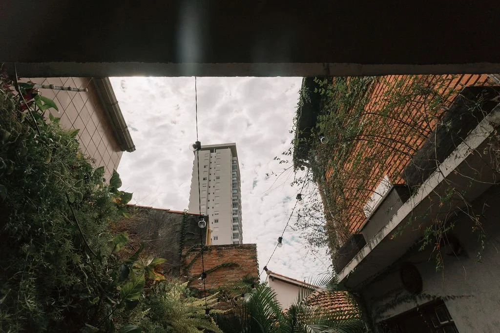 Visão de um espaço entre edifícios com plantas e uma corda de luzes decorativas, céu nublado ao fundo.