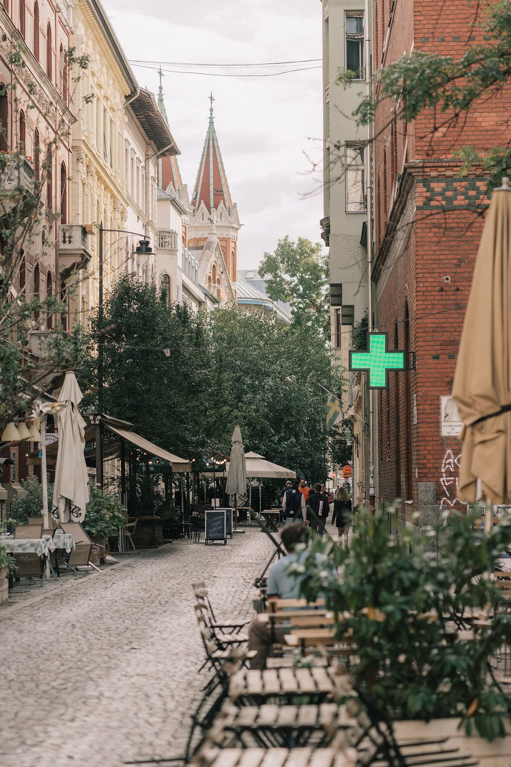 Rua de calçadão com mesas de restaurante, árvores e edifícios de vários estilos, incluindo igreja com torre, no centro da imagem, com pessoas caminhando e um símbolo de farmácia iluminado de verde na parede.