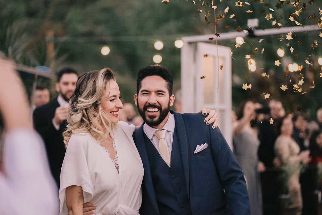 Homem e mulher em celebração de casamento, sorrindo e abraçados, com convidados ao fundo e confetes dourados caindo.