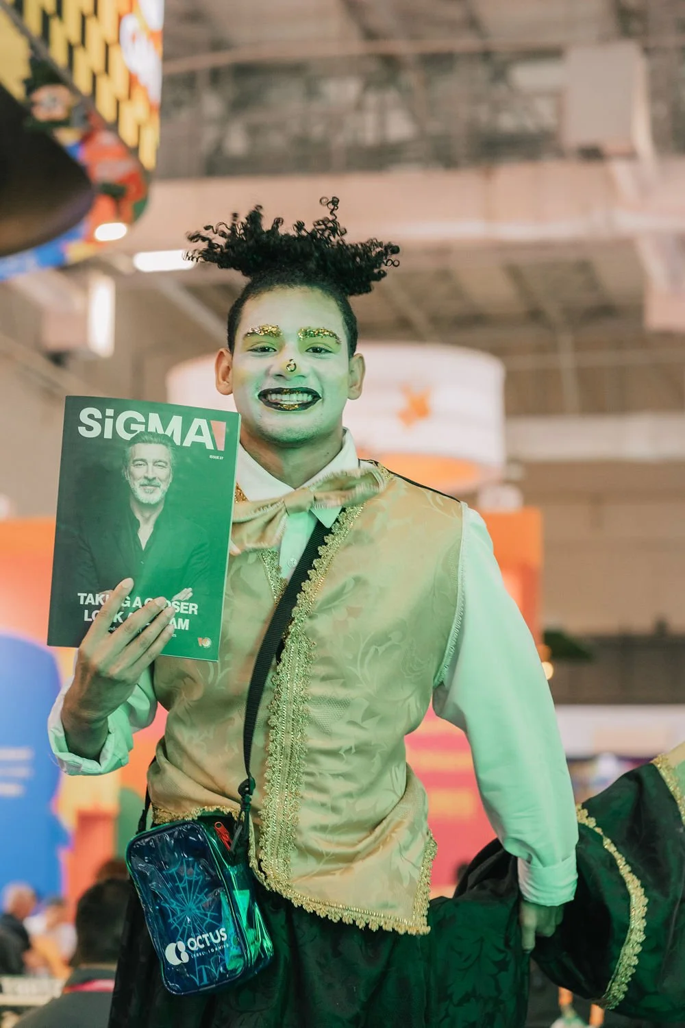 Pessoa sorridente com maquiagem e acessórios coloridos, segurando uma revista intitulada SIGMA, em um evento com fundo de iluminação brilhante.