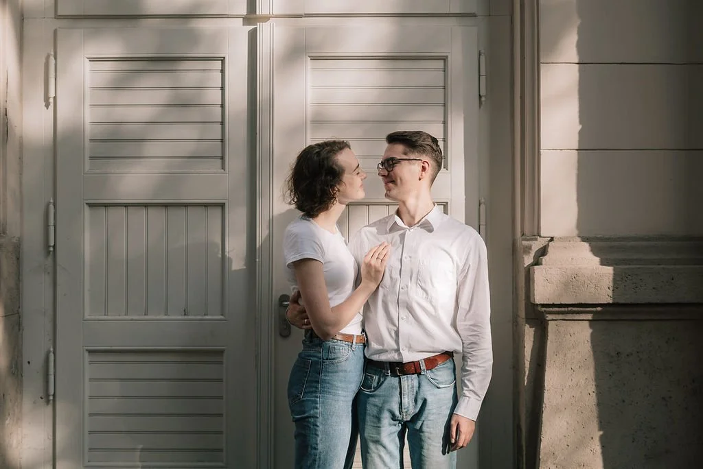 Um casal jovem em frente a uma porta branca, olhando um para o outro carinhosamente, com luz natural iluminando a cena.