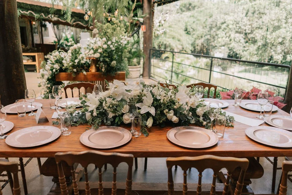 Mesa de jantar decorada com flores brancas e pratos elegantes em um ambiente ao ar livre com muita vegetação.