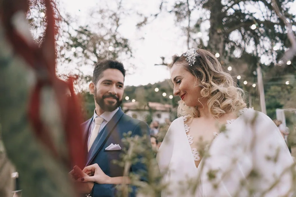 Casal de casamento ao ar livre, a noiva sorri para o noivo enquanto seguram as mãos, cenário com árvores e luzes decorativas.