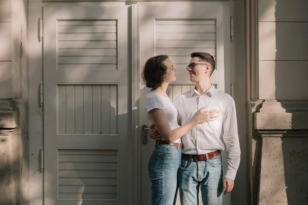 Casal sorridente olhando um para o outro na frente de uma porta branca, com paredes de pedra ao redor.