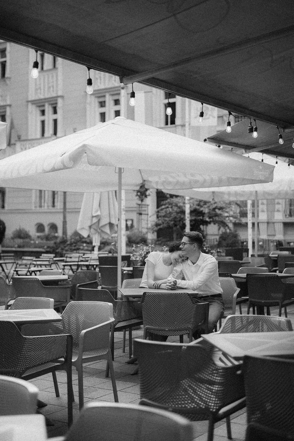 Casal sentado na mesa de um restaurante ao ar livre, compartilhando um momento romântico sob sombra de guarda-sol, com luzes decorativas penduradas acima.