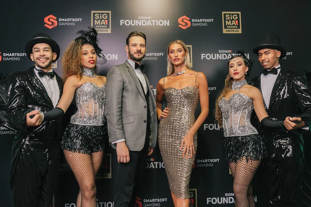 Grupo de pessoas em traje de gala posando em tapete vermelho com fundo de Logos de eventos e marcas.