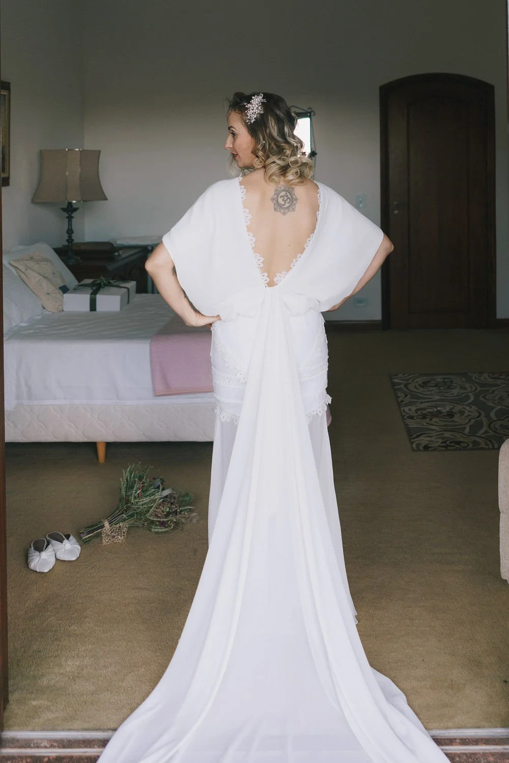 Mulher vestida de branco, de costas, com vestido de noiva aberto nas costas, mostrando uma tatuagem e um penteado com acessório de cabelo, no interior de um quarto.