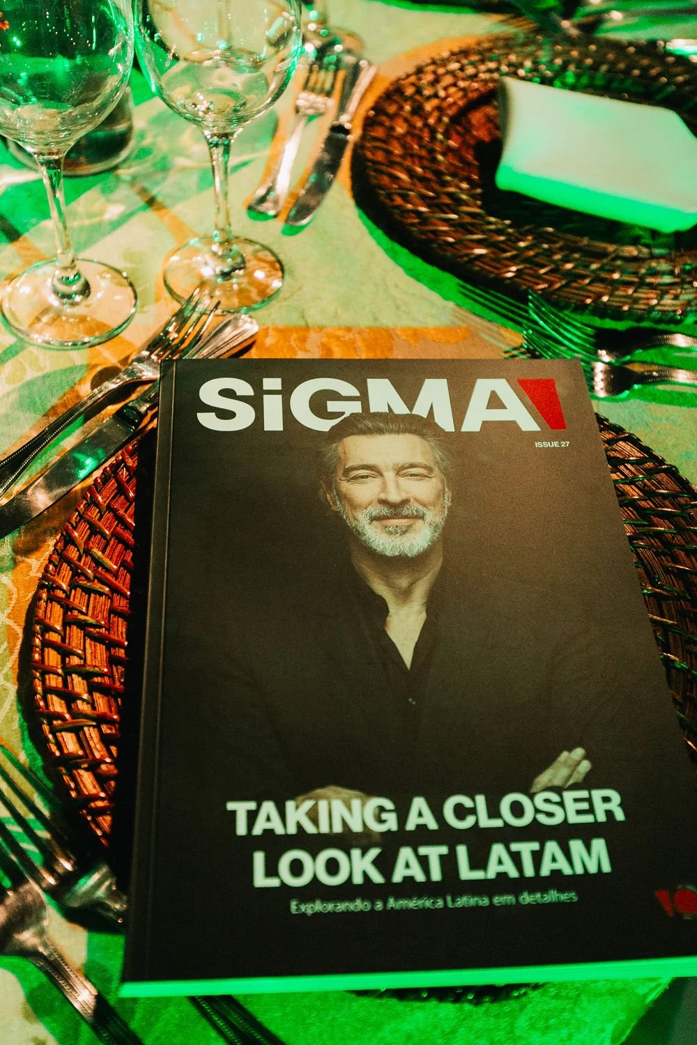 Uma mesa posta com dois copos de vinho vazios, talheres e um livro chamado 'SIGMA' com a imagem de um homem de cabelos grisalhos e barba sorrindo na capa. A mesa tem um tecido de mesa colorido e um prato de revestimento de vime ao lado.