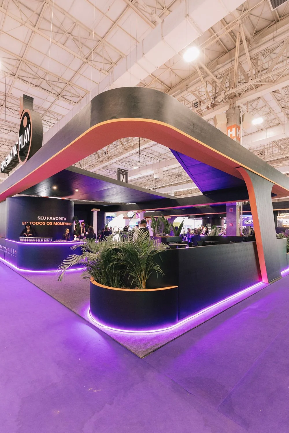Quiosque de uma feira ou evento com estrutura moderna, preta com detalhes em laranja e iluminação roxa, com plantas decorativas e várias pessoas ao redor, e um balcão que oferece bebidas.