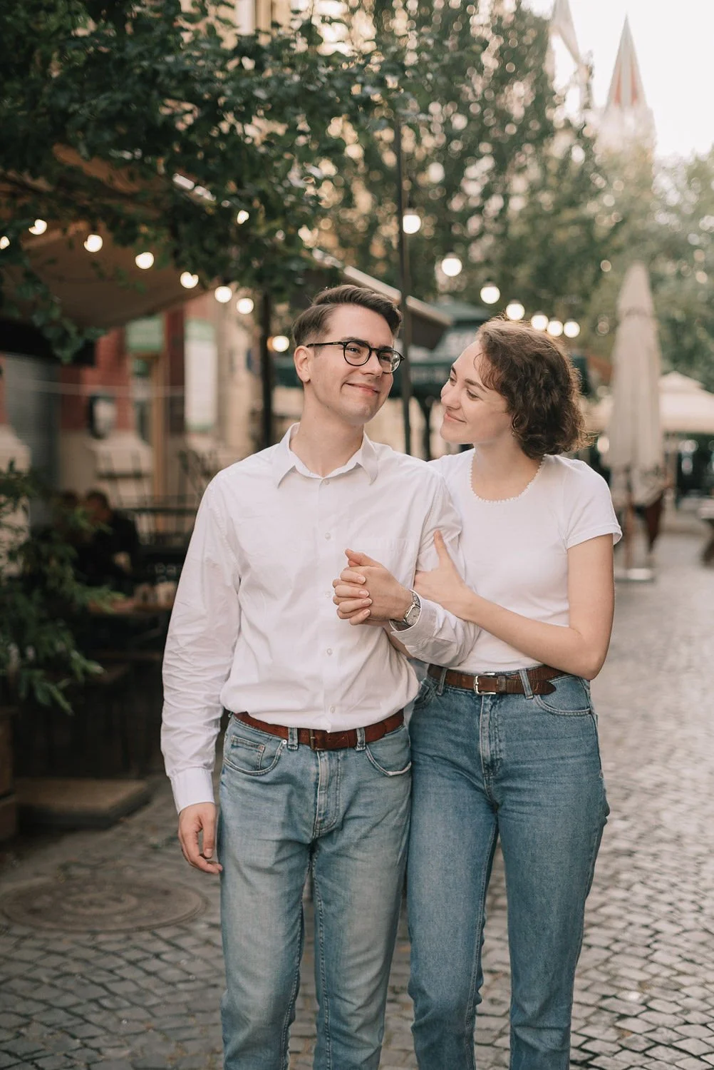 Casal caminhando de mãos dadas na rua, sorrindo e olhando um para o outro, com iluminação de luzes decorativas ao fundo.