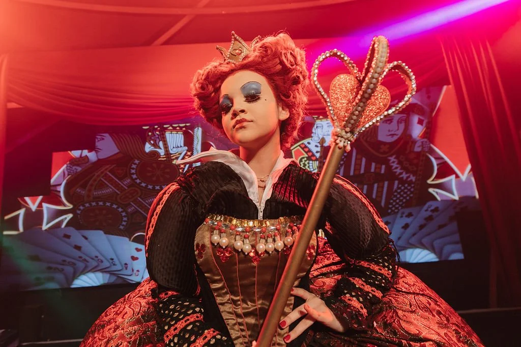Pessoa com maquiagem de rainha ou bruxa, vestindo traje elaboradamente decorado com pedras, pérolas e renda, segurando uma varinha com coração brilhante, em cenário teatral com cortinas vermelhas e fundo de imagens de cartas de baralho.