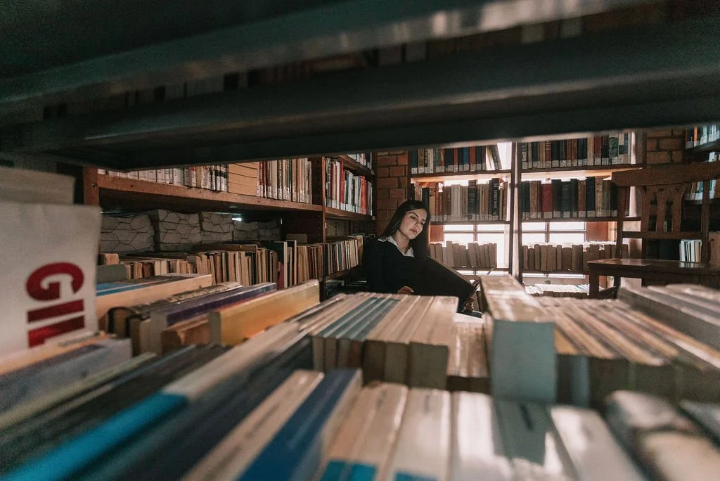 Pessoa sentada entre prateleiras de livros em uma biblioteca, vista através de uma moldura de prateleira.