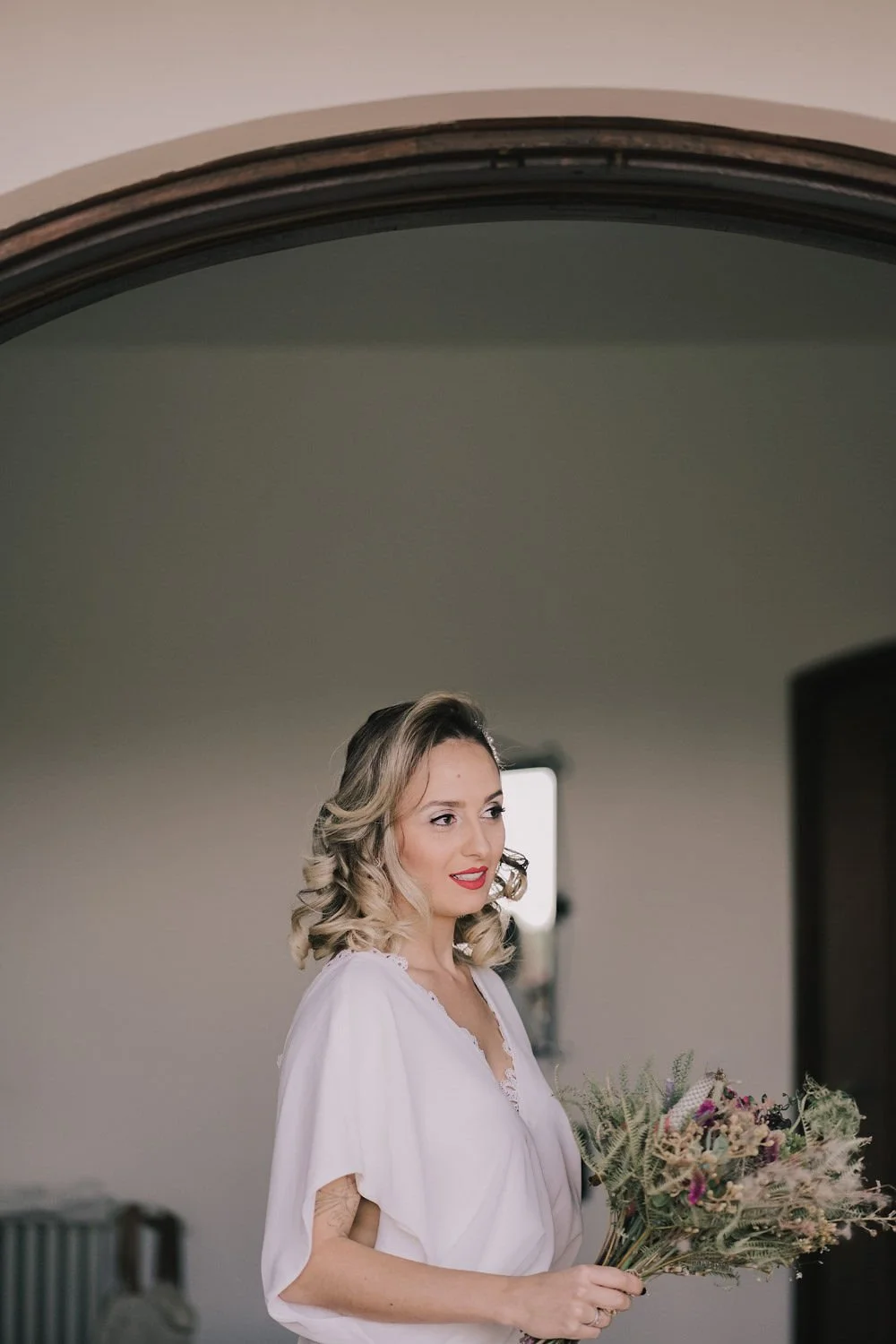 Mulher com cabelo loiro cacheado segurando um buquê de flores, posando numa parede de fundo neutro.
