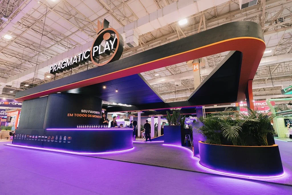 Estande de feira da Pragmatic Play com design moderno e iluminação em LED, localizado em um espaço interno de feira, com pessoas ao redor e plantas decorativas na base do estande.
