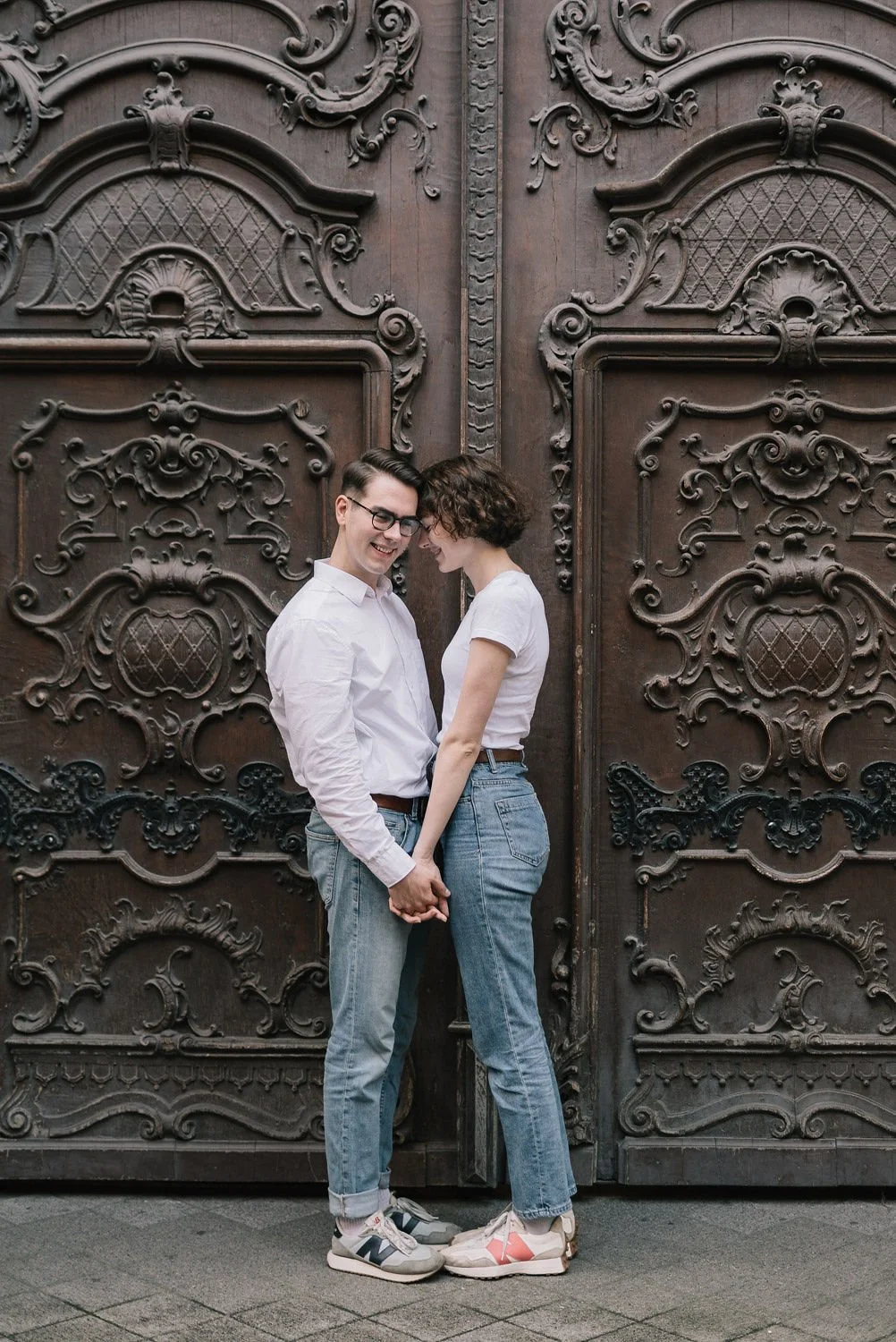 Jovens casais abraçados e sorrindo na frente de uma porta de madeira com detalhes ornamentais.