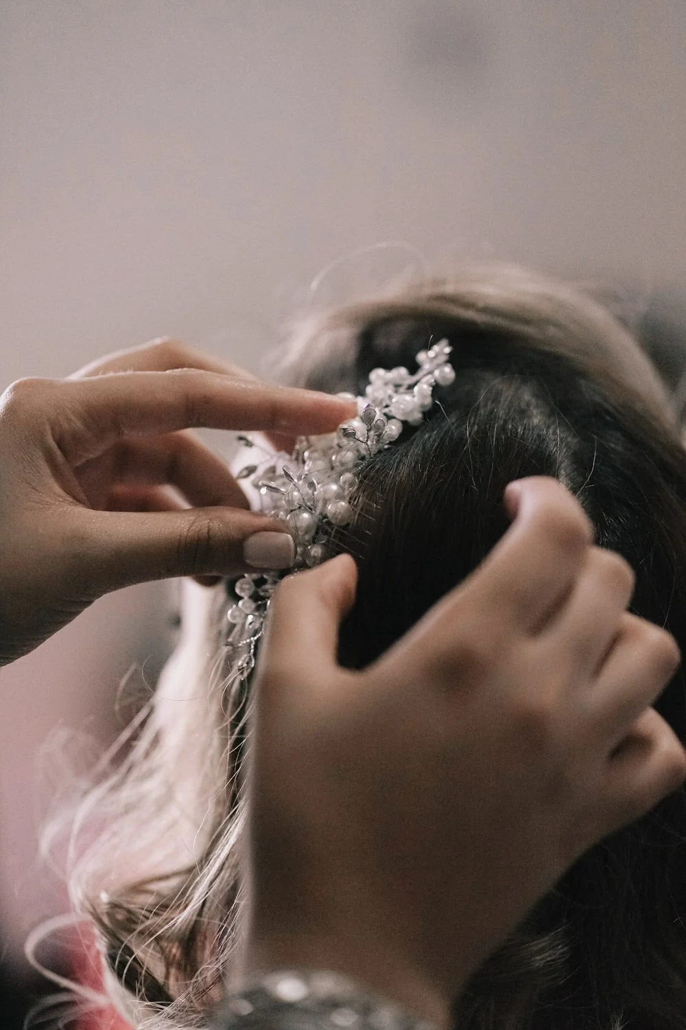 Pessoa ajustando uma tiara de pérolas e detalhes em fita e cristal na cabeça de uma mulher com cabelo escuro e loiro.