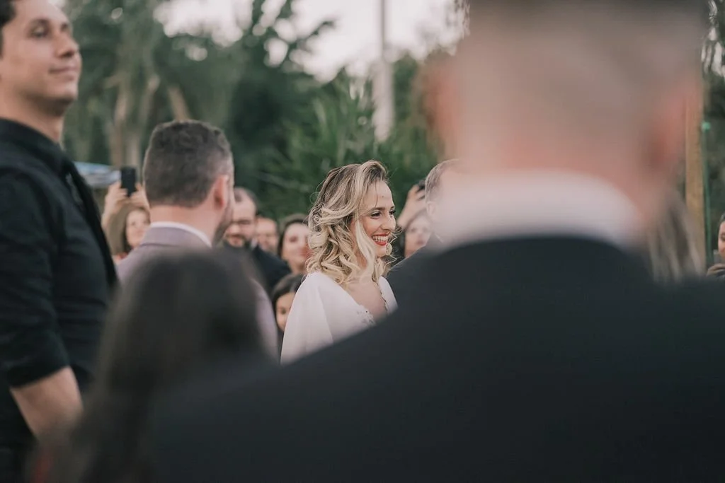 Mulher sorrindo em um evento ao ar livre com várias pessoas ao redor, roupas formais, árvores ao fundo.