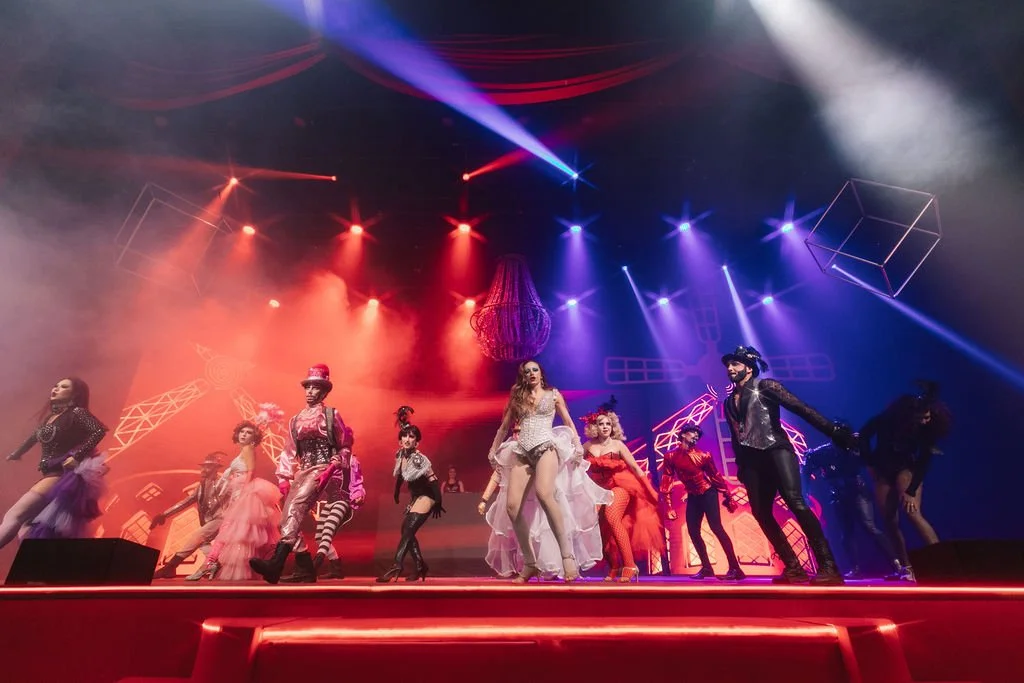 Grupo de performers em um palco com iluminação colorida, incluindo luzes vermelhas, azuis e roxas, durante uma apresentação teatral ou de dança.