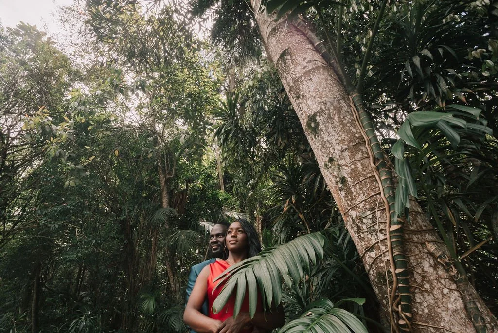 Um casal sorridente em uma floresta tropical densa, cercado por muitas plantas verdes, árvores altas e folhagens.